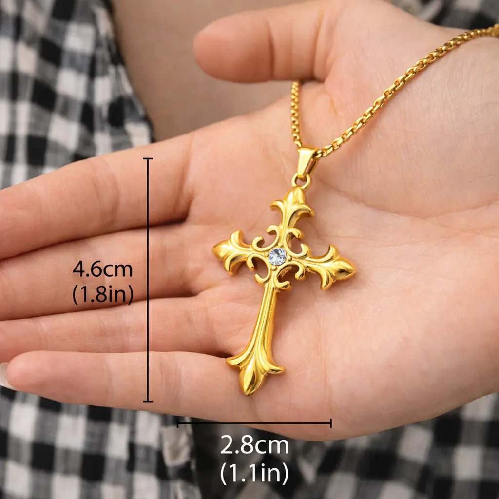 Colar Cruz Áurea™ dourado com pingente de cruz em aço inoxidável apoiado na mão, mostrando tamanho real de 4,6 cm por 2,8 cm e acabamento premium