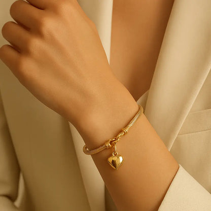 Pulseira feminina dourada com pingente de coração em aço inoxidável, usada no pulso, com design elegante e acabamento premium.